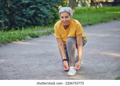 Elderly middle-aged Asia woman in sportive wear ties shoelaces ready for morning jogging or sportive walk stroll in summer park smile look at camera. Healthy active lifestyle of retiree, work out - Powered by Shutterstock