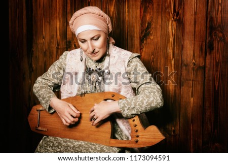 Elderly native woman in a national costume plays the ancient musical instrument.