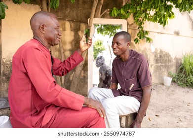 An elderly Nigerian man admires his fresh haircut in a hand mirror, smiling proudly, while his street barber observes with satisfaction, capturing personal care and traditional grooming in an outdoor  - Powered by Shutterstock