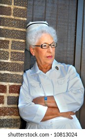 Elderly Nurse Wears Her Nursing Uniform Including Hat. She Leans Against The Brick Wall Of Old Hospital.