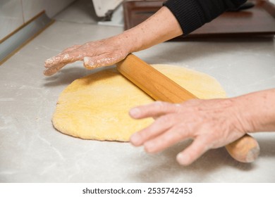 An elderly old woman grandmother rolls out the dough on a kitchen board table, hands covered in flour, close up, the dough is suitable for pizza pie bread. Food concept. - Powered by Shutterstock