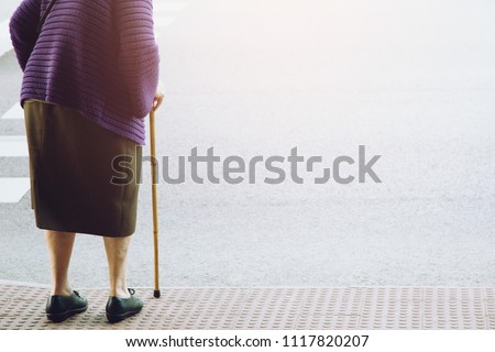 elderly old woman with walking stick stand waiting on footpath sidewalk crossing the street alone. concept senior across the street to zebra crosswalk.