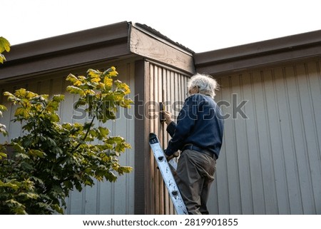 Elderly on an extension ladder using a stud finder installing new wood siding boards, homeowner maintenance and upkeep, making outdoor repairs on house
