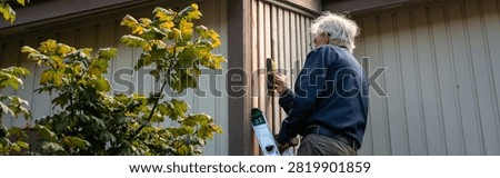 Elderly on an extension ladder using a stud finder installing new wood siding boards, homeowner maintenance and upkeep, making outdoor repairs on house
