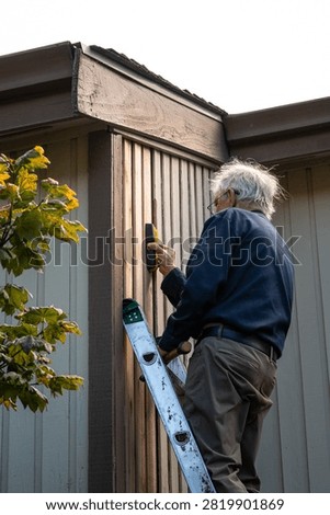 Elderly on an extension ladder using a stud finder installing new wood siding boards, homeowner maintenance and upkeep, making outdoor repairs on house
