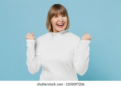Elderly overjoyed happy woman 50s wearing white knitted sweater doing winner gesture celebrate clenching fists say yes isolated on plain blue color background studio portrait. People lifestyle concept - Shutterstock ID 2105097206