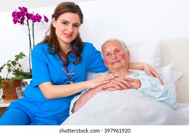 Elderly Patient Laying In Bed In A Nursing Home With A Caring Young Female Doctor.