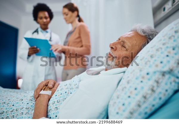 Elderly\
patient recovering in the hospital ward while his daughter is\
communicating with a doctor in the\
background.