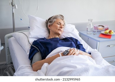 Elderly patient sleeping on a medical bed in hospital ward. Senior woman resting after operation with eyes closed and IV drip. Old mature woman lying on bed and wearing blue hospital gown. - Powered by Shutterstock