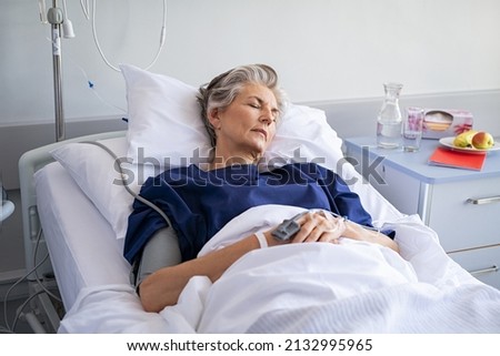 Elderly patient sleeping on a medical bed in hospital ward. Senior woman resting after operation with eyes closed and IV drip. Old mature woman lying on bed and wearing blue hospital gown.