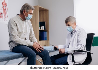 Elderly patient wearing medical mask talking to male family therapist during regular doctors appointment. Senior man, suffering knee pain, taking care of his health during coronavirus pandemic. - Powered by Shutterstock
