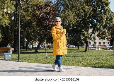 Elderly people and healthy lifestyle concept. Senior woman in casual clothes and fashionable glasses holding cup of coffee or tea, enjoying walk in park outdoors on sunny day, looking at the camera.