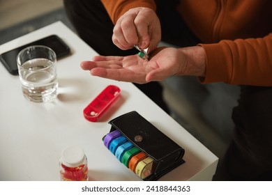 Elderly person taking pill into hand, with water glass, smartphone, and pill organizer in the background. Daily vitamins and meds. Concept of health and medical care, aging, medicine, treatment - Powered by Shutterstock