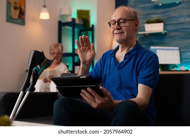 Elderly Person Waving At Tablet With Video Call Conference On Sofa With Crutches. Old Man Using Online Remote Communication While Senior Woman Sitting In Wheelchair Reading