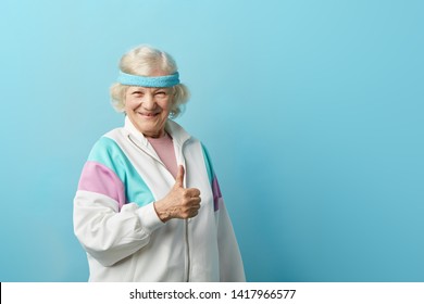 Elderly positive woman in sports wear and headband giving a thumb up and looking at the camera isolated on blue background