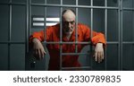 Elderly prisoner in orange uniform holds hands on metal bars, looks at camera. Criminal serves term of imprisonment in prison cell. Inmate stands behind bars in jail or detention center. Portrait.