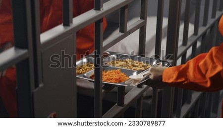 Elderly prisoner in orange uniform sits in prison cell. Prison guard gives him tray of food through metal bars. Guilty criminal serves imprisonment term for crime. Inmate in jail or detention center.