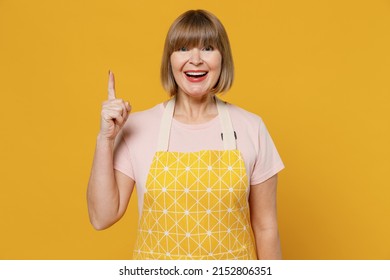 Elderly proactive fun insighted housekeeper housewife woman in orange apron holding index finger up with great new idea isolated plain on yellow background studio. People household lifestyle concept