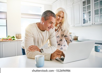 Elderly Retired Couple Reading Their Social Media News On A Laptop Computer In A Fresh White Kitchen As They Enjoy A Relaxing Retirement