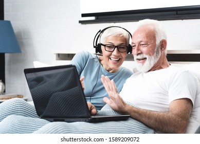 Elderly retired couple talking over skype with their son who living far away over the ocean. Senior woman has in-ear headphones with a microphone. Concept maintaining distance contact.