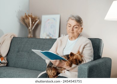 Elderly retired senior woman with wrinkles smiling while embracing her Yorkshire terrier dog pet and relaxing while reading on sofa at home. Best friend. Enjoying retirement lifestyle. - Powered by Shutterstock