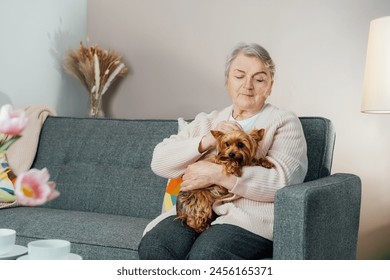 Elderly retired senior woman with wrinkles smiling while embracing her Yorkshire terriers dog pet and relaxing with pet on sofa at home. Best friend. Enjoying retirement lifestyle. - Powered by Shutterstock