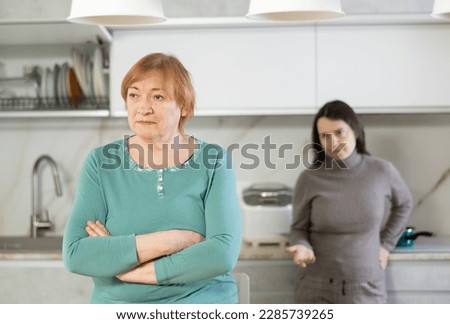 Elderly sad woman sits at table in kitchen and listens to insults.Daughter-in-law stands behind elderly lady and expresses her indignation.