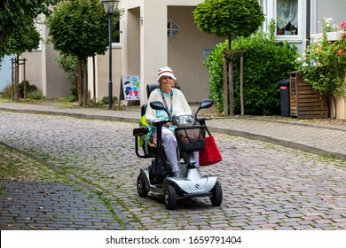 An Elderly Satisfied Lady Rides An Electric Scooter Along The Street Of The Old German City. Image Of Active Old Age, A Taste For Life, Vitality In Old Age.. Bad Wimpfen, Germany - August 8, 2019