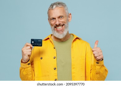 Elderly Satisfied Smiling Happy Fun Gray-haired Mustache Bearded Man 50s Wear Yellow Shirt Hold In Hand Credit Bank Card Show Thumb Up Isolated On Plain Pastel Light Blue Background Studio Portrait.