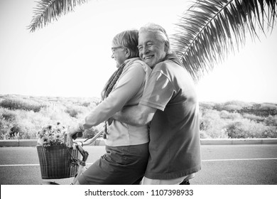 Elderly Senior Caucasian Couple Play And Enjoy Leisure Activity Outdoor In Lifestyle Vacation. Man And Woman Retired Go On A Vintage Bike Like Children Age. Tropical Palm Background.