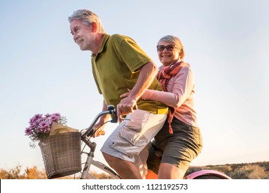 Elderly Senior Caucasian Couple Play And Enjoy Leisure Activity Outdoor In Lifestyle Vacation. Man And Woman Retired Go On A Vintage Bike Like Children Age. Tropical Palm Background.