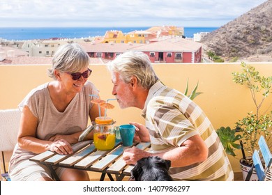 Elderly Senior Couple Man And Woman With Nice Dog Pug On The Terrace. Drink Together From The Same Vase A Fruit Juice For Healthy Lifestyle. Summer Nice Time Of Vacation In A Terrace And Ocean View