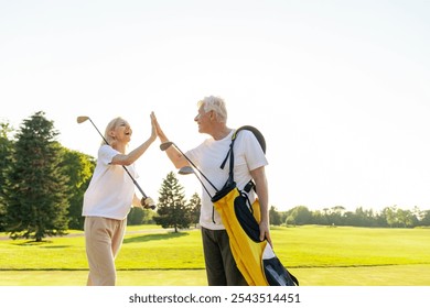 elderly senior couple in uniform celebrating victory and success in golf game and giving high five, old man and woman playing golf on golf course at sunset and doing outdoor sports - Powered by Shutterstock