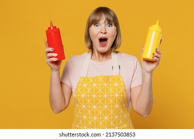 Elderly Shocked Surprised Happy Housekeeper Housewife Woman 50s Wearing Orange Apron Hold Ketchup And Mustard Isolated Plain On Yellow Background Studio Portrait. People Household Lifestyle Concept