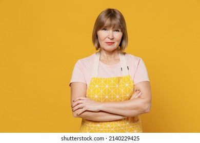 Elderly Shrewd Calm Serious Caucasian Housekeeper Housewife Woman Wearing Orange Apron Hold Hands Crossed Folded Isolated Plain On Yellow Background Studio Portrait. People Household Lifestyle Concept