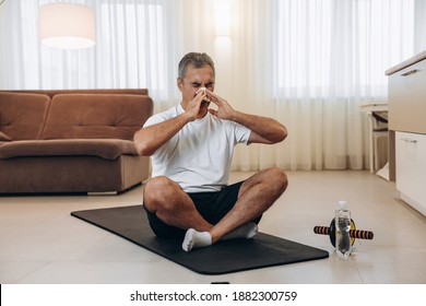 Elderly Sick Man With Runny Nose Sitting On Black Yoga Mat. Stay Home Concept. Man Blows His Nose After Workout Or Fitness. Practicing Sports At Home If You Feel Sick. Workout With Sports Equipment.