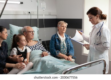 Elderly Sick Old Patient Resting In Bed With Caring Family Beside Him Recovering After Medical Surgery In Hospital Ward. Physician Doctor Woman Checking Senior Man During Clinical Therapy