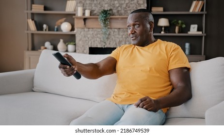 Elderly Single Bachelor African American Adult Senior Man Sitting On Couch Watching TV Enjoy Serial Movie Comments News Program On Television Use Remote Control Switch Sport Channels Relaxing At