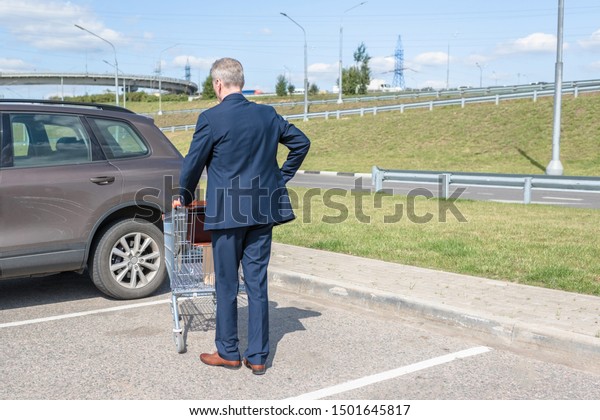 Elderly single man in a\
blue suit walks to his car with a supermarket trolley. Businessman\
and shopping