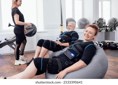 Elderly smiling couple sits on massage chairs after ems workout in gym. Young female trainer using remote control sets necessary parameters on electrical muscle stimulation suit of grayheaded man. - Powered by Shutterstock