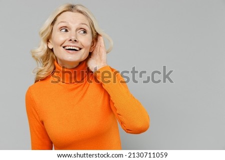 Elderly smiling fun happy blonde caucasian woman 50s in orange turtleneck try to hear you overhear listening intently isolated on plain grey color background studio portrait. People lifestyle concept