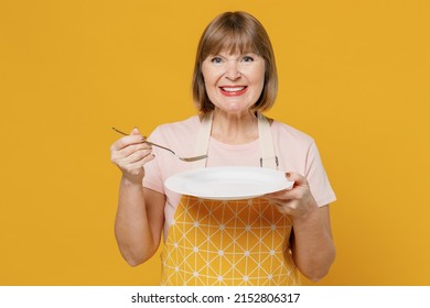 Elderly Smiling Fun Happy Cooker Housekeeper Housewife Woman 50s Wear Orange Apron Hold Empty Plate Spoon Try Meal Eat Isolated Plain On Yellow Background Studio. People Household Lifestyle Concept