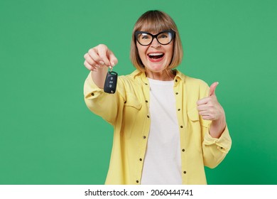 Elderly smiling happy caucasian woman 50s in glasses yellow shirt hold car keys fob keyless system show thumb up gesture isolated on plain green background studio portrait People lifestyle concept.