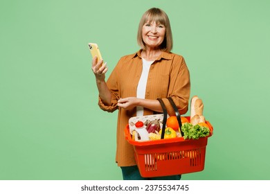 Elderly smiling happy cheerful woman wear brown shirt casual clothes hold shopping basket bag with food products use mobile cell phone isolated on plain green background. Delivery service from shop - Powered by Shutterstock