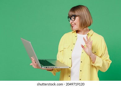 Elderly Smiling Happy Fun Caucasian Woman 50s In Glasses Yellow Shirt Hold Use Work On Laptop Pc Computer Waving Hand Isolated On Plain Green Color Background Studio Portrait People Lifestyle Concept