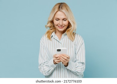 Elderly Smiling Happy Fun Caucasian Woman 50s Wearing Striped Shirt Hold In Hand Use Mobile Cell Phone Isolated On Plain Pastel Light Blue Color Background Studio Portrait. People Lifestyle Concept