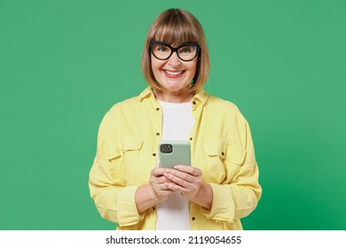 Elderly Smiling Happy Satisfied Woman 50s Wearing Glasses Yellow Shirt Hold In Hand Use Mobile Cell Phone Browsing Internet Isolated On Plain Green Background Studio Portrait People Lifestyle Concept