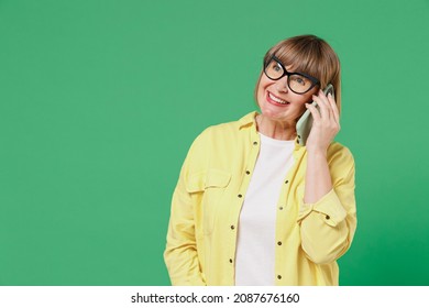 Elderly Smiling Happy Woman 50s In Glasses Yellow Shirt Talk Speak On Mobile Cell Phone Conducting Pleasant Conversation Isolated On Plain Green Background Studio Portrait People Lifestyle Concept.