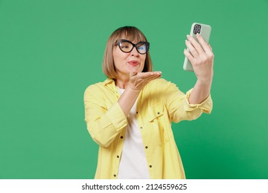 Elderly Smiling Happy Woman 50s In Glasses Yellow Shirt Doing Selfie Shot On Mobile Cell Phone Post Photo On Social Network Blowing Air Kiss Talk Video Call Isolated On Plain Green Background Studio