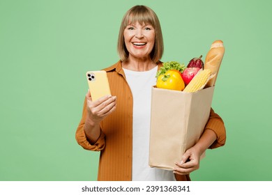 Elderly smiling happy woman wear brown shirt casual clothes hold shopping paper bag with food products hold in hand use mobile cell phone isolated on plain green background. Delivery service from shop - Powered by Shutterstock